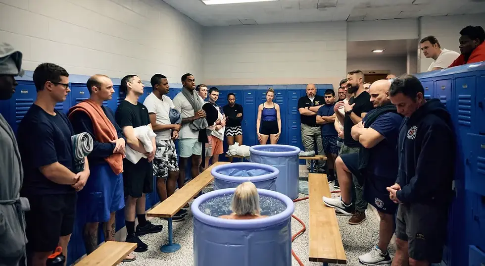 A group gathers in a locker room around three ice baths, watching someone immerse in cold water; the mood is focused and supportive.