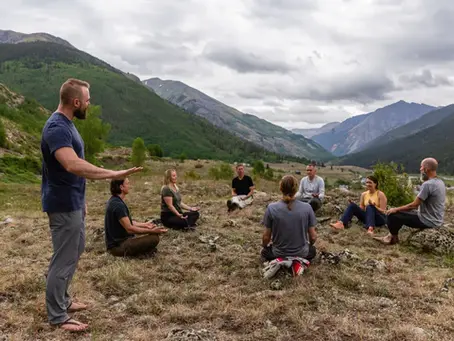 A guide gestures to a seated group on a rocky alpine meadow, surrounded by green mountains beneath a cloudy sky; the gathering feels calm and reflective.
