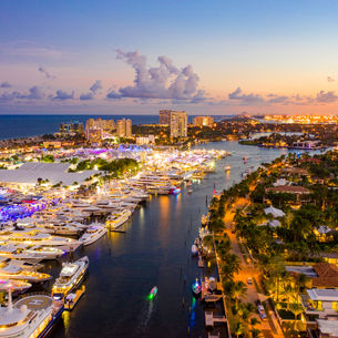 Aerial view of Fort Lauderdale's Intracoastal Waterway at dusk filled with numerous yachts and boats, illuminated by colorful lights. The scene includes a waterway lined with palm trees and luxurious homes on one side, and high-rise buildings near the beach on the other, with the ocean visible in the background under a sky with scattered clouds and a sunset glow.