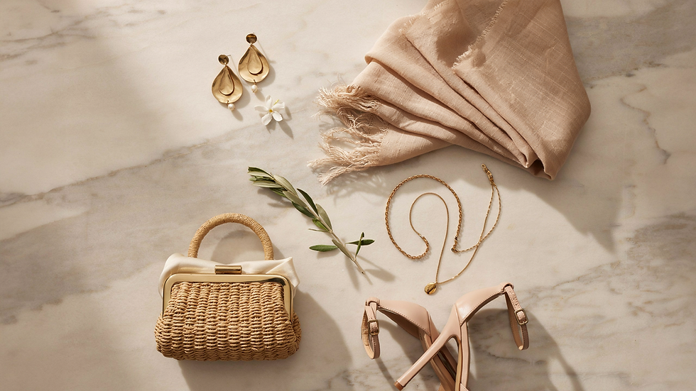 Flat lay of blush scarf, gold earrings, necklace, wicker handbag, nude heels, and olive sprig on marble, warm and elegant.