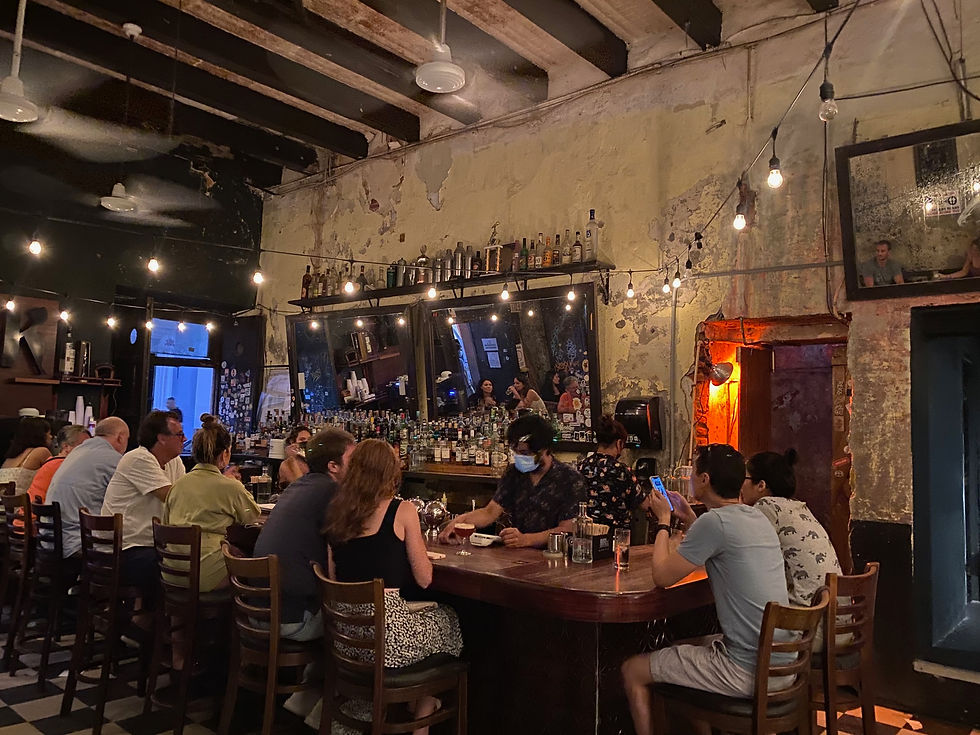 Dimly lit bar with patrons seated at a wooden counter. Bartender serves drinks. Worn walls, string lights, and bottles are visible. Cozy atmosphere.