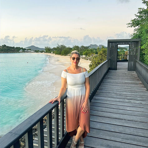 Woman in a gradient dress stands on a boardwalk by the beach at sunset, with turquoise water and distant greenery. Calm and serene mood.