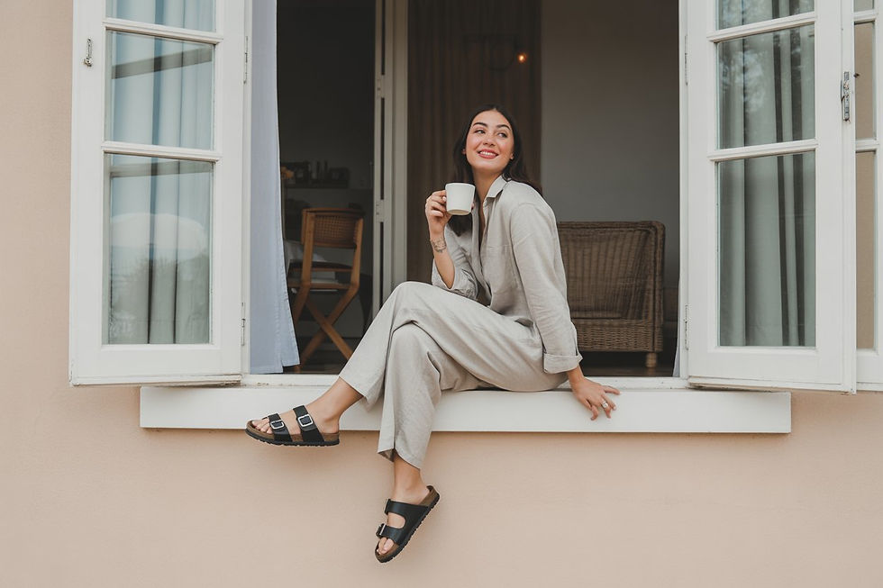 Woman in beige loungewear sits in a windowsill, smiling and holding a mug. Neutral exterior, open white windows, relaxed mood.