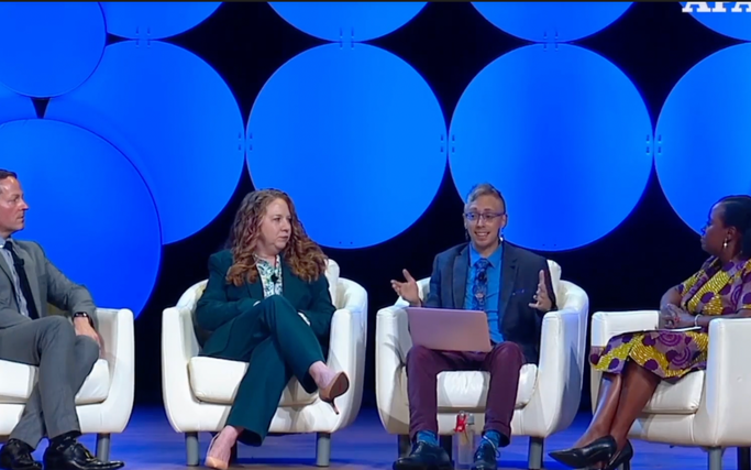 4 panelists sit in a semi-circle, three panelists dressed professionally (a white cis man, a white cis woman, and a Black cis woman) looking towards Dr. Kris Gebhard (a white genderqueer transmasculine person dressed in a blue blazer, lighter blue shirt and darker blue tie with blue earrings), who is gesturing and appears to be speaking.]
Dr. Gebhard provides consultation on gender-affirming care and building community resilience within organizations and groups that are resisting state-sanctioned violence.