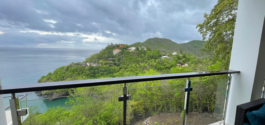 A hotel balcony with a scenic view, featuring a small table and two chairs, surrounded by lush greenery and blue skies.