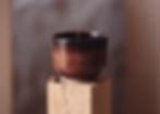 Front view of a modern, minimalist, ceramic Espresso cup with brown and red shaded glaze on wood with a coffee bean.