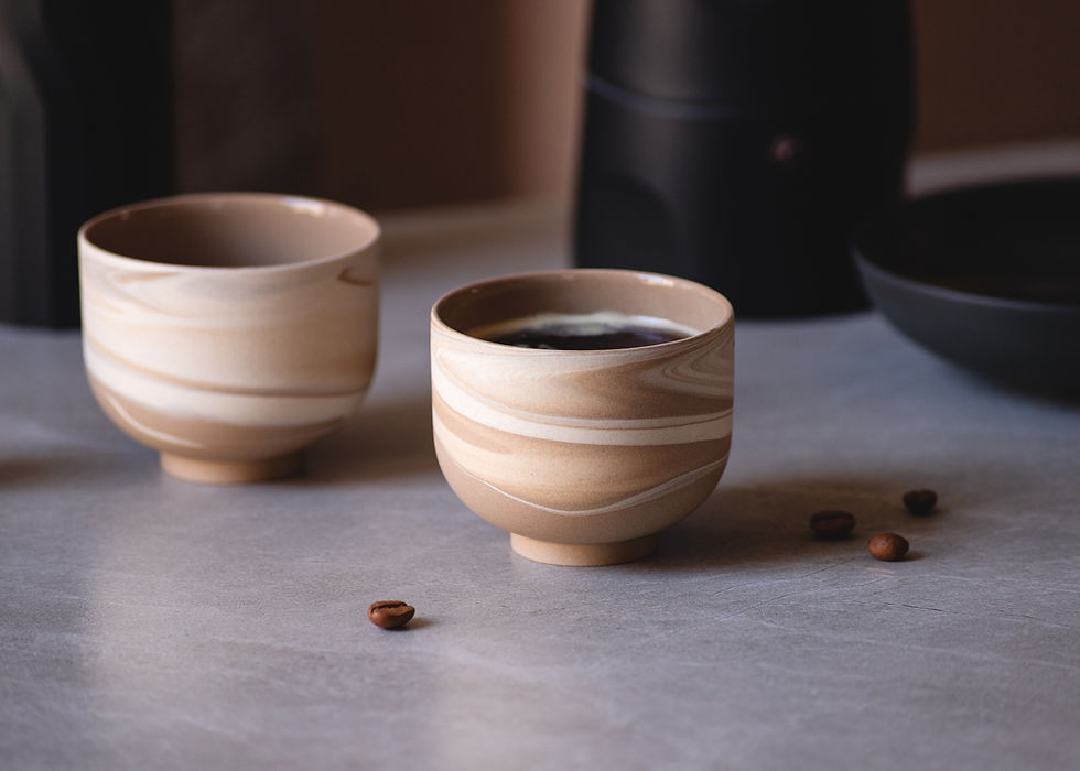 Thumbnail: Close up of two minimalist, round Cortado cups in matte brown with white swirls with coffee on a white marble surface.