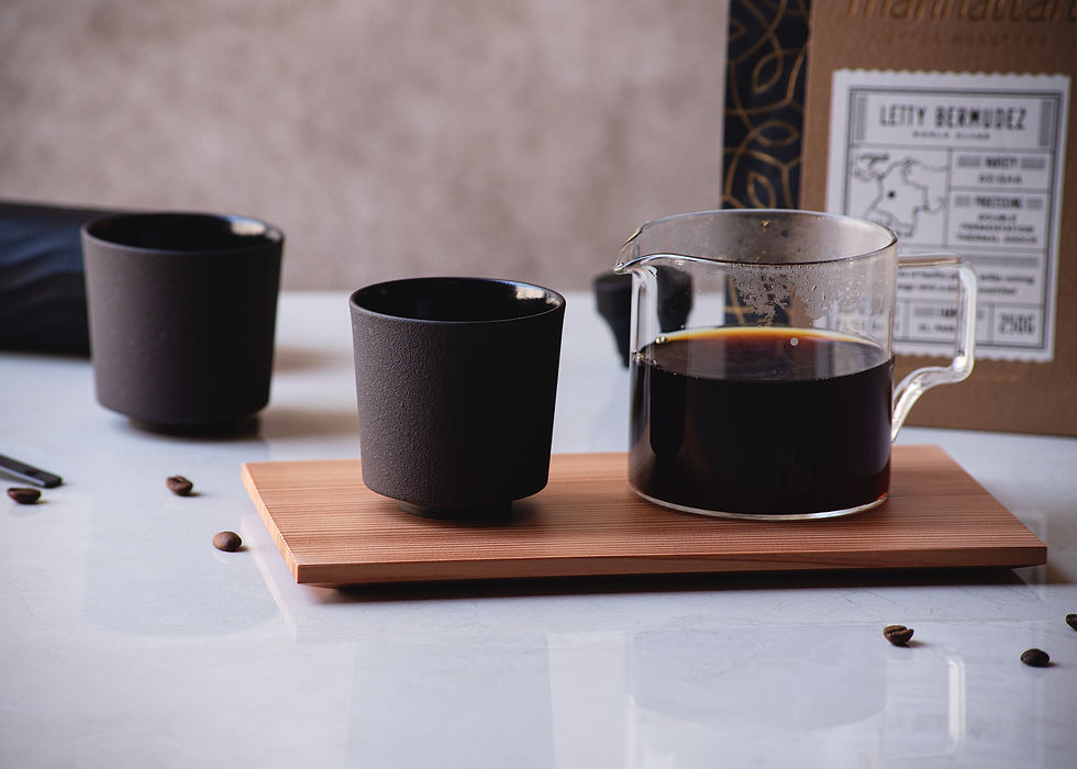 A wooden tray holds a glass coffee carafe and two dark ceramic cups, set on a marble table with coffee beans scattered around