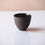 Thumbnail: A small, black, textured ceramic cup sits on a smooth, light-colored surface with a soft shadow below it.