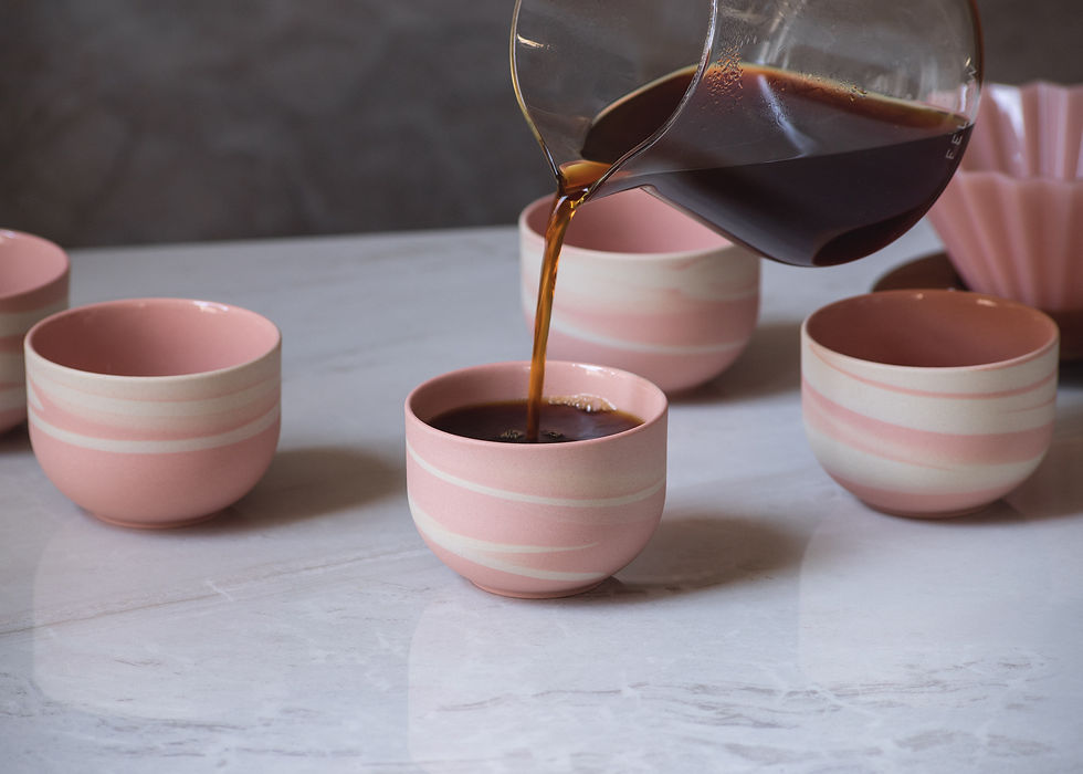 Thumbnail: Front view of three minimalist, round, ceramic cup in pink with white swirls. Coffee is pouring from glass vessel into one.