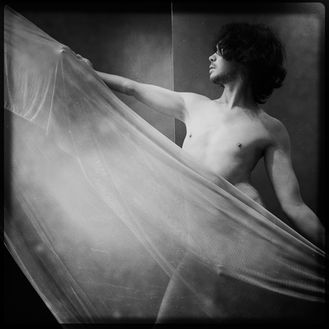 black and white photo of a dark haired nude male twisting and holding up a curtain