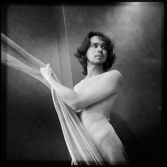black and white photo of a dark haired nude male clutching a curtain and turning to face behind himself