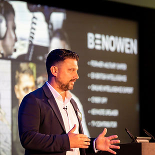 Ben Owen speaking at an event, gesturing in front of a screen that showcases his background in military intelligence and operational expertise, with a slideshow of images in the background.