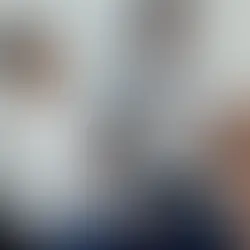 Man in a white shirt smiles, holding a tablet and handing a document to a woman. Another woman in a suit watches. Bright, indoor setting.