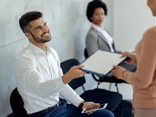 Man in a white shirt smiles, holding a tablet and handing a document to a woman. Another woman in a suit watches. Bright, indoor setting.