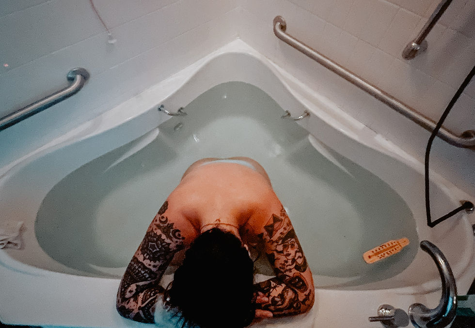 Tattooed person in a corner bathtub, head resting on arms. The tub has handle bars, water is calm, creating a serene mood.