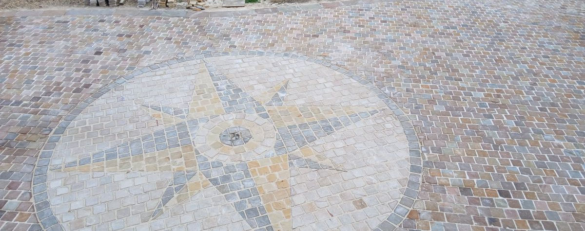 Vue aérienne détaillée d'un pavage complexe en grès multicolore, centré sur une rose des vents géométrique précise, avec une zone de chantier et des meubles de jardin en arrière-plan.