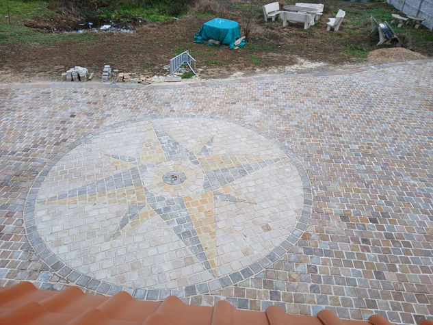 Vue aérienne détaillée d'un pavage complexe en grès multicolore, centré sur une rose des vents géométrique précise, avec une zone de chantier et des meubles de jardin en arrière-plan.
