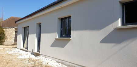 Vue d'ensemble de la façade latérale d'une maison neuve ravalée en beige clair, montrant trois fenêtres à menuiserie sombre et l'ombre nette d'un arbre sur la structure.