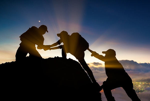 Three people hiking on a hill. One is holding the hand of the guy at the center so he can get up and the third one is pushing the center guy so he can climbed,