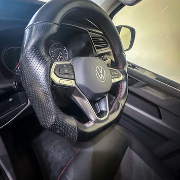 Close-up interior view of a Volkswagen camper van’s driver area, focusing on a leather-trimmed multifunction steering wheel with perforated grips and contrast stitching. Dashboard controls, instrument cluster, and door panel details are visible, showcasing a modern cabin designed for comfort, convenience, and everyday driving.