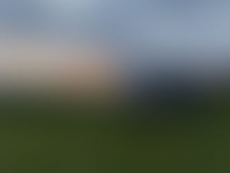 A silver SC Sporthomes van is parked on a grassy coastal site, facing the ocean as the sun sets on the horizon. The scene is bathed in warm, golden-orange light, with the sun partially hidden by clouds, casting a soft glow across the sky and reflecting off the rippling water. Low hedges line the edge of the grass, separating the van from the beach below. A second campervan is faintly visible in the distance to the right, emphasizing the peaceful, communal nature of this coastal retreat. The atmosphere is serene and cinematic, capturing a perfect moment at the end of a road trip day—where comfort, mobility, and nature converge in a stunning, seaside setting.