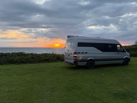 A silver SC Sporthomes van is parked on a grassy coastal site, facing the ocean as the sun sets on the horizon. The scene is bathed in warm, golden-orange light, with the sun partially hidden by clouds, casting a soft glow across the sky and reflecting off the rippling water. Low hedges line the edge of the grass, separating the van from the beach below. A second campervan is faintly visible in the distance to the right, emphasizing the peaceful, communal nature of this coastal retreat. The atmosphere is serene and cinematic, capturing a perfect moment at the end of a road trip day—where comfort, mobility, and nature converge in a stunning, seaside setting.