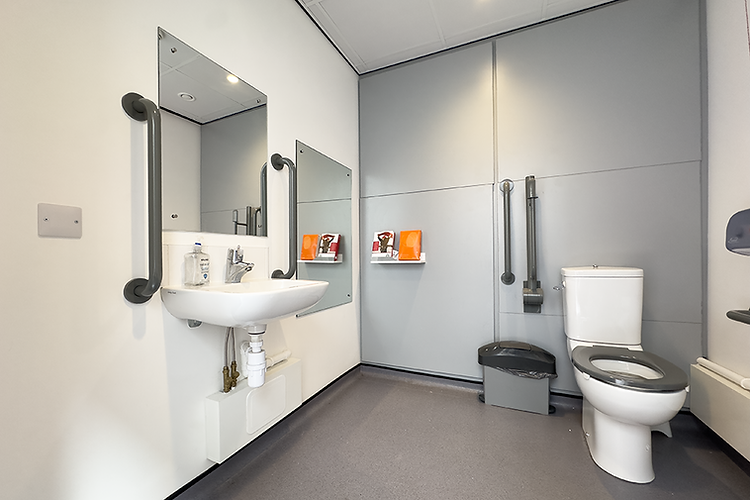Interior of an accessible public restroom designed for wheelchair users. The room features a white sink with exposed plumbing and two vertical grab bars on either side, mounted beneath a large mirror. A standard-height toilet with a black seat is positioned on the right side, next to a waste bin and a vertical support rail attached to the wall. The walls are a combination of white and light grey panels, with a shelf holding sanitary products such as pads and tampons. The floor has a non-slip grey finish, and the overall space is clean, modern, and compliant with accessibility standards.