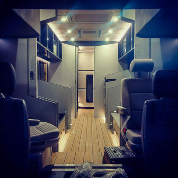 Looking down the central aisle of a luxury motorhome conversion, showcasing a finished interior with recessed lighting, wooden plank flooring, and sleek cabinetry. The view leads toward a futuristic rounded pod feature at the rear. Overhead compartments, modern wall panels, and clean lines highlight the advanced design and craftsmanship typical of high-end, custom RV builds.