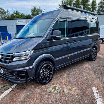 Front three-quarter view of a grey VW camper van with RJ Howard Bespoke Conversions branding and Avondale Motorpark decals. It features black multi-spoke alloy wheels, a roof-mounted awning, and privacy-tinted windows. The van is parked in an industrial area with other vehicles and commercial units in the background.