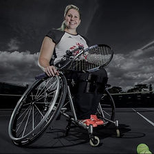 A positive and empowering female wheelchair tennis player on an outdoor court, holding a racket on her lap with a determined smile. She wears a graphic tee and sports shorts, complemented by vibrant orange sneakers, against a dramatic cloudy sky. A tennis ball is beside her chair on the court.