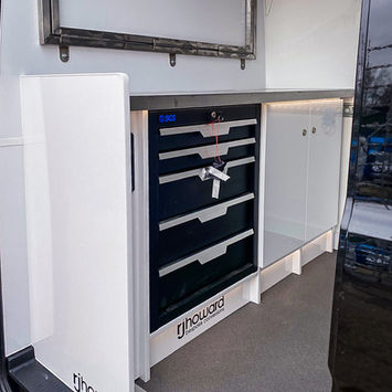 Rear-side view of the workstation area inside the van. A custom-fitted SGS tool chest is built into white gloss cabinetry with a stainless steel countertop above. RJ Howard branding is printed along the lower panel and cabinet side wall.