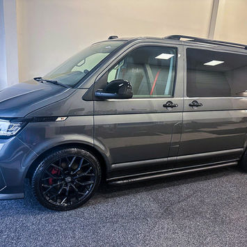 Side profile view of a metallic gray Volkswagen camper van displayed inside a conversion workshop. The van features black multi-spoke alloy wheels with red brake calipers, black side steps, roof rails, privacy glass, and a sleek modern body kit. Through the front window, premium seats with contrasting red seatbelts are visible, highlighting the vehicle’s custom camper conversion and sporty styling.