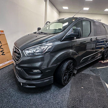 Front three-quarter view of a dark gray Ford Transit custom van displayed inside a workshop. The van features a metallic paint finish, black alloy wheels, tinted windows, side steps, and a sporty body kit that enhances its modern appearance. Large headlights, a bold front grille, and sculpted body lines give the vehicle a refined yet purposeful look. Positioned on a workshop floor with conversion and detailing equipment nearby, the van showcases a premium custom build designed for practicality, style, and everyday usability.