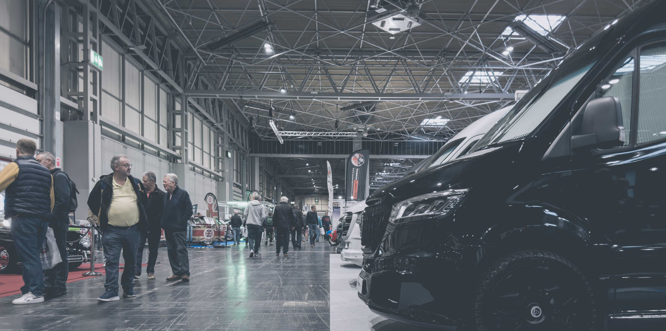 Visitors explore a busy indoor motorhome exhibition where a range of premium camper vans are displayed beneath the expansive steel roof of a modern exhibition hall. In the foreground, the front corner of a sleek black camper van dominates the frame, while attendees walk between exhibition stands and engage with displays throughout the spacious venue. Bright overhead lighting and wide aisles create an open atmosphere, highlighting the event as a destination for discovering the latest recreational vehicles, touring innovations, and luxury travel solutions.
