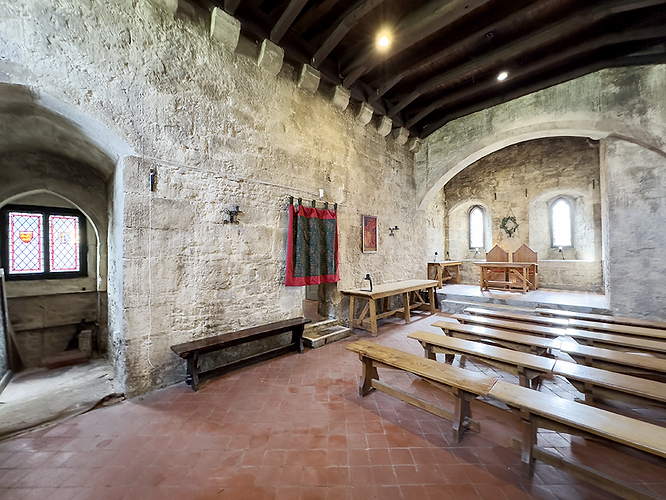 A historic stone hall with high arched ceilings and exposed wooden beams, lit by natural light streaming through narrow stained-glass and arched windows. The room features long wooden benches arranged in rows facing a raised platform at the far end, where two carved wooden chairs sit beneath the arched window. Against the side walls are simple wooden tables and benches, while a red-and-green tapestry hangs near the center. The terracotta tiled floor and thick, aged stone walls give the space a medieval atmosphere, suggesting it was once used as a dining or gathering hall within a castle or fortress.