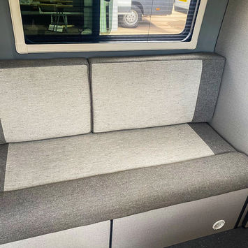 Close-up of a light grey upholstered bench seat inside a camper van, with a window above reflecting a view of a van and workshop area. The clean design features squared cushions and minimalist styling, set against a light and modern interior wall.