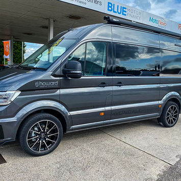 Angled side view of a dark grey camper van with RJ Howard Bespoke Conversions branding and stylish black alloy wheels. The vehicle is parked at a motor dealership forecourt beneath signs for Blue Motor Finance and RAC Warranty. A roof-mounted Fiamma awning is fitted, and the van has tinted windows and a sporty body kit.