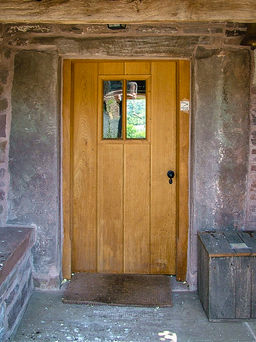 A solid oak front door is centered within a weathered stone wall, framed by thick stone pillars and a rustic timber beam overhead. The door is vertically panelled and features a small, square window at eye level, divided into four panes. To the right is a vintage iron door handle, and a wooden bench sits nearby. The ground outside the door is rough concrete with a brown doormat in front.