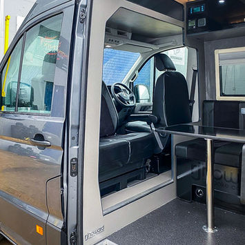 Side door open view of a camper van revealing the driver’s cab and a compact dining area inside. The black leather seat bench with integrated seatbelts sits across from a central metal pedestal table. The flooring is finished in a durable speckled vinyl, and the interior walls are clad in light grey panels with black accents. The "rjhoward" logo is visible on the door frame, indicating a bespoke conversion by the company.