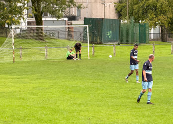 A goalkeeper in a green jersey sits injured by the goalpost, with the referee bending down to check on him, while teammates walk away concerned.