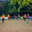 Players from both teams in yellow-and-black and navy-and-light-blue walk across the pitch during a pause in play, with leafy trees and pitch-side adverts in the background.
