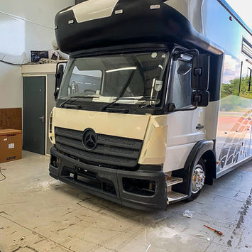 Workshop image of a large, custom motorhome with a black and cream exterior. The front bumper and lighting elements are still being installed, and protective wrapping is visible on the body. The over-cab sleeping area extends forward in a curved, aerodynamic design. The vehicle sits on shiny chrome wheels inside a clean, industrial garage space equipped for vehicle fabrication.