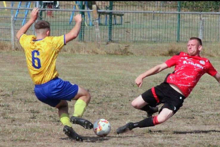 Two football players contesting for the ball on a dry, patchy grass pitch. A player in a yellow jersey with the number 6 and blue shorts attempts to control the ball while raising his arms, as an opposing player in a red and black kit slides in aggressively to challenge. The ball is positioned between them, and the intensity of the tackle is clear. A metal fence and playground equipment are visible in the blurred background.
