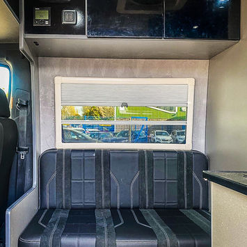Bench seat inside a camper van with black leather upholstery and vertical stitching, positioned beneath a wide window with a closed blind and overhead black storage cabinets.