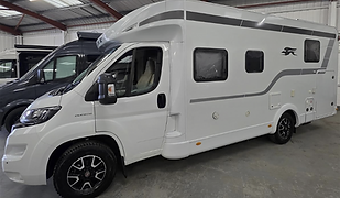 A white Fiat Ducato-based motorhome is parked indoors on a smooth concrete floor, with other camper vans visible in the background of the warehouse-style setting. The motorhome has a clean, modern profile with subtle grey graphics, including a swooping stripe and a stylized bird logo on the rear half. It features large side windows, black alloy wheels, and multiple access hatches for utilities and storage along the side. The overhead cab area extends slightly forward, suggesting extra sleeping space inside. Bright lighting from the corrugated metal roof above highlights the vehicle’s sleek contours, making this an ideal display setting for showcasing the practicality and design of a compact yet capable touring motorhome.