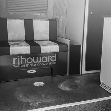 Black-and-white image of a camper van's interior showing a two-tone bench seat labeled “rjhoward bespoke conversions.” A round floor mount is visible in front, and the flooring shows visible swirls and marks, possibly from cleaning or wear. The area is softly lit, with upper cabinets and a window blind in view.