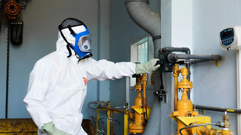 A worker wearing a full white protective suit, blue full-face respirator mask, and green gloves adjusts equipment in an industrial setting. The worker is interacting with a control mechanism connected to yellow piping and valves, surrounded by additional metallic pipes and gauges. A wall-mounted digital display nearby shows readings, indicating a monitored environment. Overhead, industrial components such as a pulley system are visible, emphasizing a controlled and technical workspace. The scene conveys precision, safety, and specialised operations.