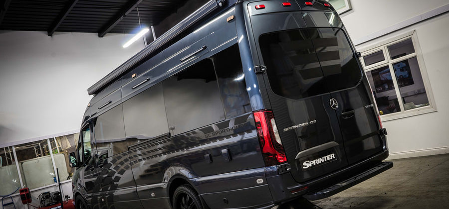 The rear view of a luxury Mercedes-Benz Sprinter van is captured indoors, showcasing its sleek design. The glossy dark gray body reflects surrounding lights, while tinted rear windows and red LED taillights add to its modern aesthetic. Black alloy wheels complement the vehicle’s polished look, and subtle decals enhance its side panels. The spacious van is equipped with a roof-mounted awning, offering versatility for outdoor use. The industrial setting highlights the van’s high-end, professional finish.
