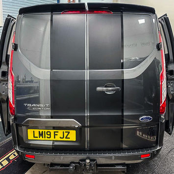 Rear view of a dark gray Ford Transit Custom van with both rear doors opened outward. Black racing stripes run vertically across the rear doors, complementing the sporty appearance of the vehicle. A tow hitch is mounted beneath the bumper, while tall vertical taillights frame the rear opening. The registration plate and Transit Custom badging are clearly visible, and the workshop setting highlights the vehicle’s customized presentation. The design blends commercial utility with distinctive styling details for a unique, professional look.